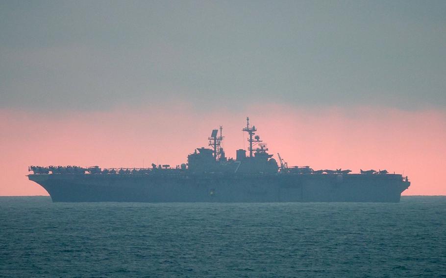 A U.S. Navy amphibious assault ship is silhouetted against a dramatic sunset sky with pink and orange hues. 