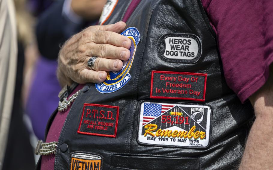 Close-up view of the torso of a man wearing a black leather vest covered with patches that have slogans written on them. The man has his hand over his heart.