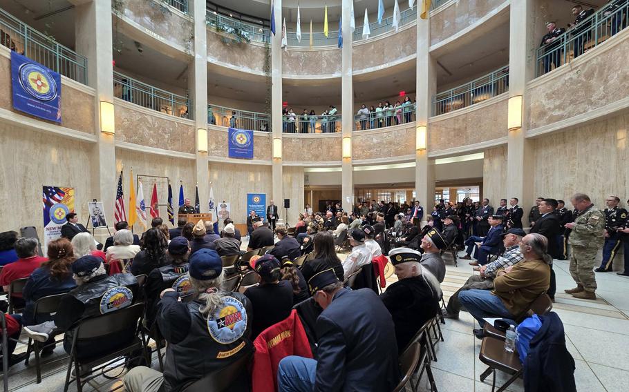 A wide-angle photo of a rotunda inside a government building filled with people seated in chairs and spectators standing at a railing on the floor above.