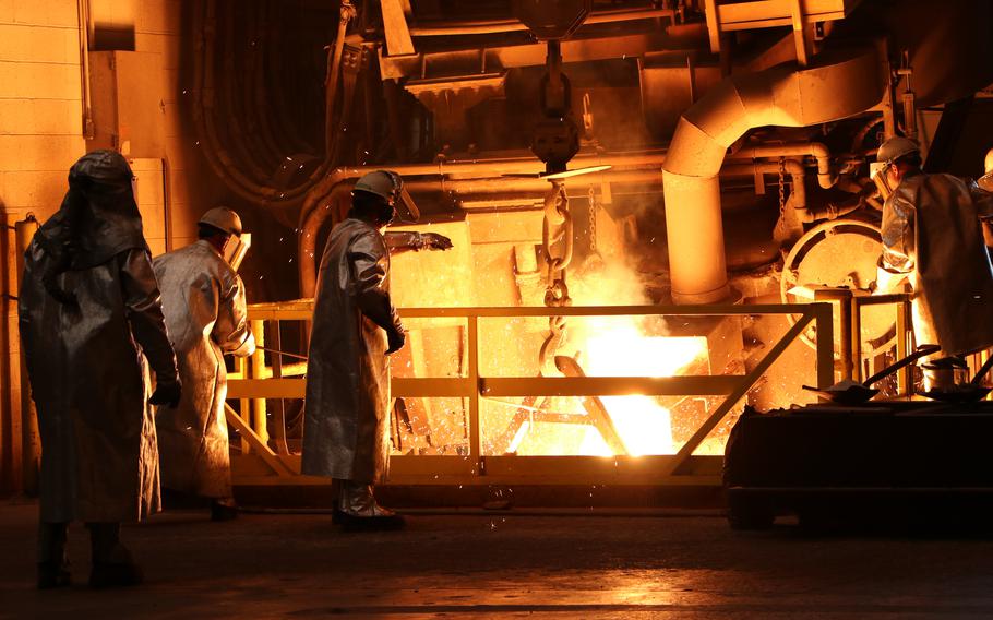 The fire of a foundry illuminates several people in protective gear.
