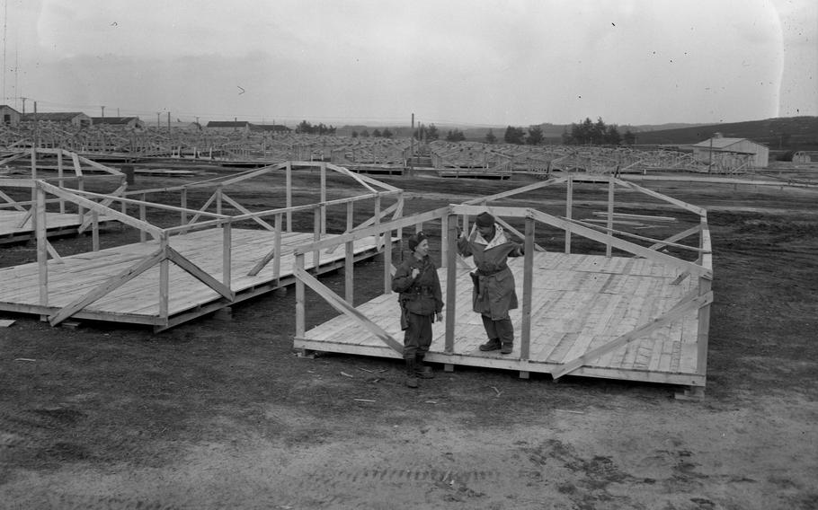 Two soldiers talk and smile while standing near the foundational structures for wooden barracks.