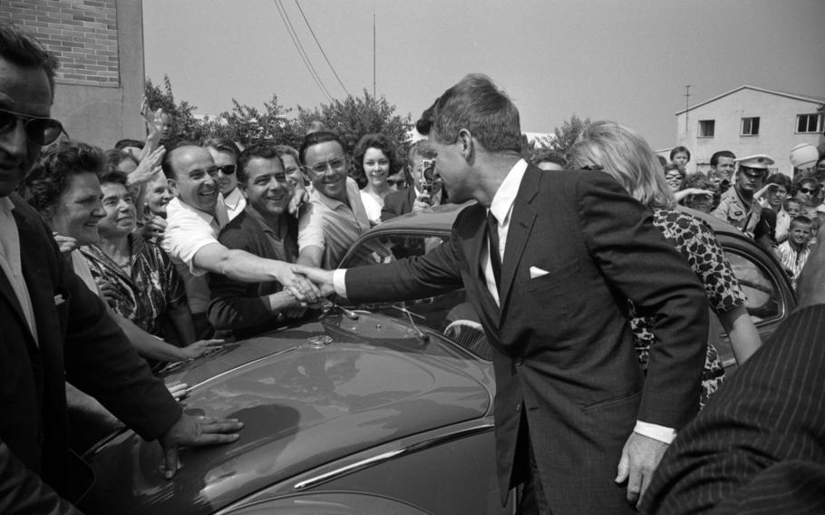 RFK reaches across his car’s hood to shake someone’s hand as fans surround him.