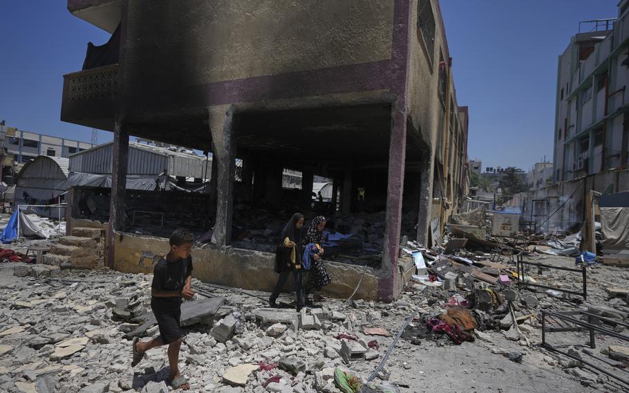 Children walk past a partially destroyed building, on a street littered with rubble.