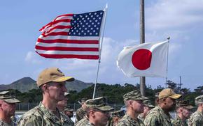 U.S. and Japanese flags fly above the heads of military service members in camouflage uniforms and hats standing at attention.