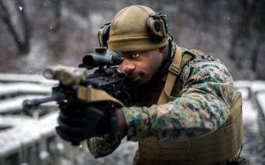 A marine in camouflage uniform and tactical gear aims a rifle with an optical scope in a snowy outdoor environment. The person is wearing a tan helmet with mounted equipment, protective eyewear and a tactical vest.