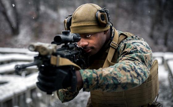 A marine in camouflage uniform and tactical gear aims a rifle with an optical scope in a snowy outdoor environment. The person is wearing a tan helmet with mounted equipment, protective eyewear and a tactical vest.