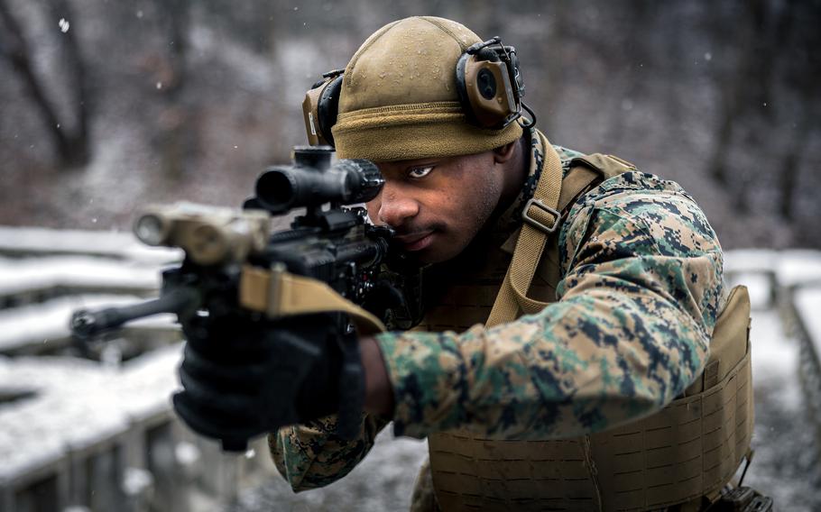 A marine in camouflage uniform and tactical gear aims a rifle with an optical scope in a snowy outdoor environment. The person is wearing a tan helmet with mounted equipment, protective eyewear and a tactical vest. .