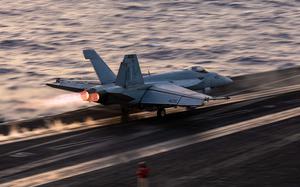 A military fighter jet speeds down the flight line as it takes off from an aircraft carrier at sea.