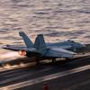 A military fighter jet speeds down the flight line as it takes off from an aircraft carrier at sea.