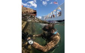 A diver in scuba gear is seen just below the surface of water next to the wreckage of a ship, with the sky above the waterline at the top of the scene.