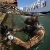 A diver in scuba gear is seen just below the surface of water next to the wreckage of a ship, with the sky above the waterline at the top of the scene.