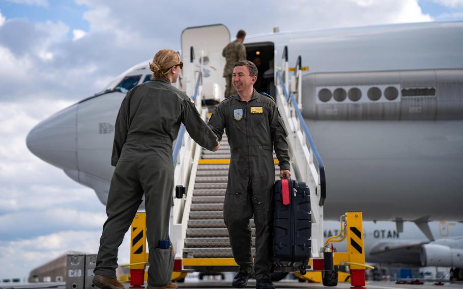 Two military personnel in flight suits are seen on an airport tarmac near the open stairs of a large aircraft. One individual, carrying a suitcase, is descending the stairs and smiling while shaking hands with the other individual at the bottom. 