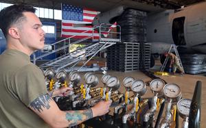 A military serviceman in an olive green T-shirt operates a control panel with multiple pressure gauges and yellow valve handles during aircraft load testing. He has tattoos on his forearms and is focused on adjusting the controls.