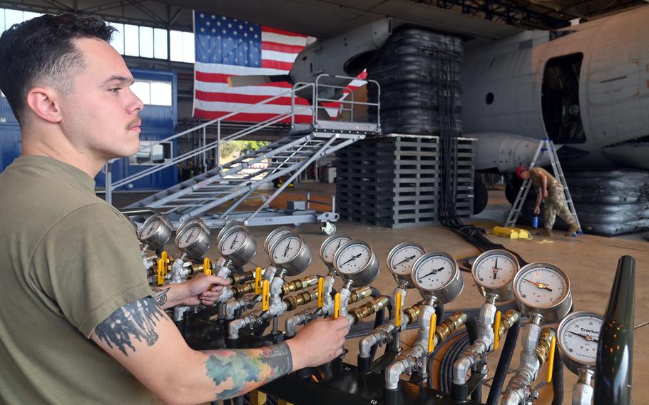 A military serviceman in an olive green T-shirt operates a control panel with multiple pressure gauges and yellow valve handles during aircraft load testing. He has tattoos on his forearms and is focused on adjusting the controls.