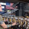A military serviceman in an olive green T-shirt operates a control panel with multiple pressure gauges and yellow valve handles during aircraft load testing. He has tattoos on his forearms and is focused on adjusting the controls.