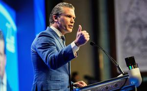Pete Hegseth, seen from the side, gestures with his hand as he stands at a podium on a stage and speaks into a microphone.