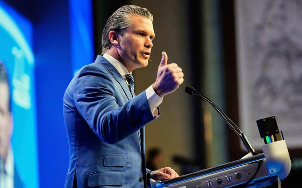 Pete Hegseth, seen from the side, gestures with his hand as he stands at a podium on a stage and speaks into a microphone.