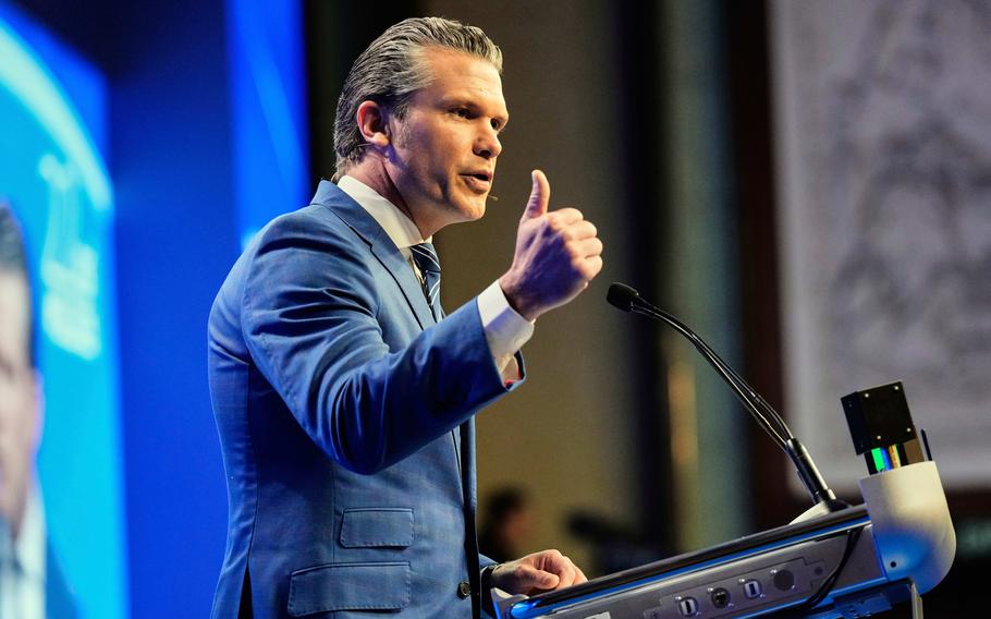 Pete Hegseth, seen from the side, gestures with his hand as he stands at a podium on a stage and speaks into a microphone.