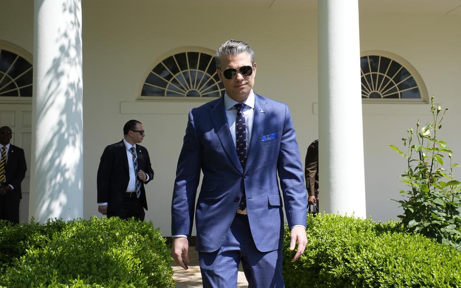 Defense Secretary Pete Hegseth wearing a suit and sunglasses walks outside.
