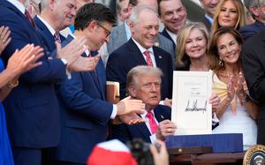 Trump, with members of congress and other onlookers applauding, holds up the law showing his signature.