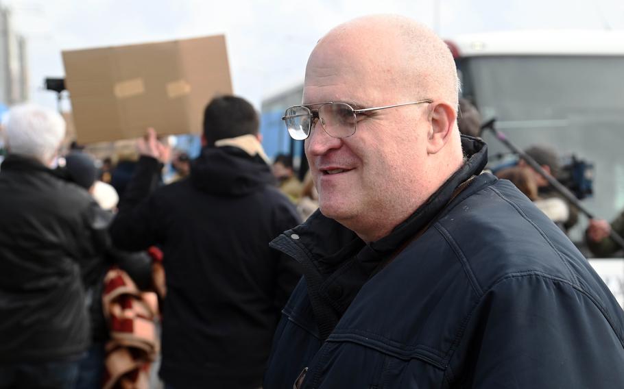 Raymond Heckard, an American who was living with his family in Ukraine, steps off a bus at a transit site for refugees near the border crossing at Korczowa, Poland, Feb. 27, 2022. In the background, people hold up signs offering rides to places in Poland, Lithuania, Latvia and the Czech Republic, to the refugees arriving.