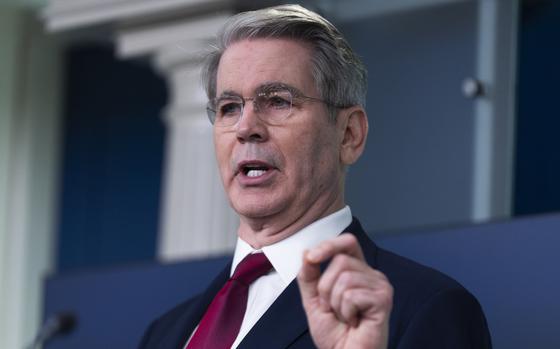 A man in a dark suit and red tie, seen from the chest up, gestures with his left hand while speaking into a microphone.