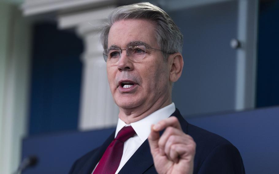 A man in a dark suit and red tie, seen from the chest up, gestures with his left hand while speaking into a microphone.