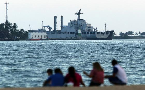 People seen out-of-focus sitting near a body of water, with an in-focus warship farther away ahead of them.