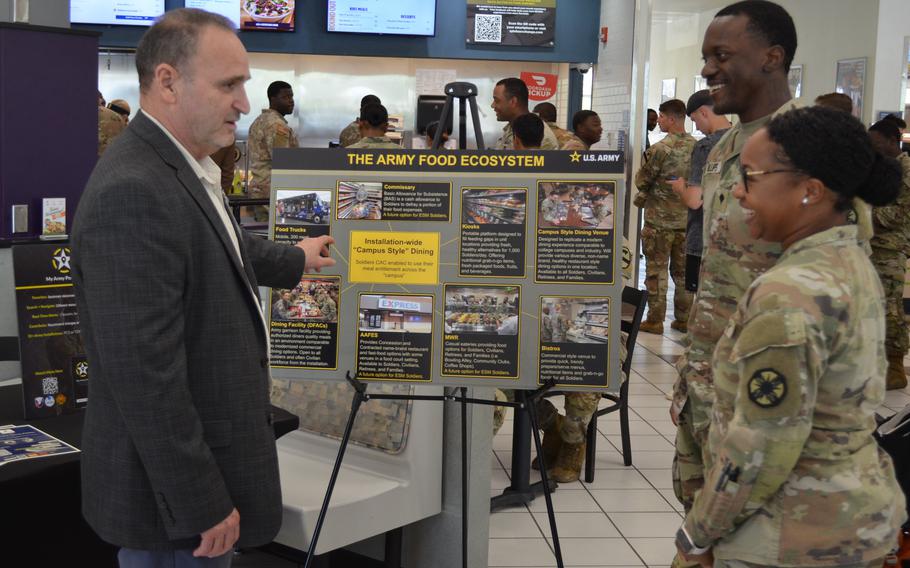Two soldiers chat with a man who is pointing at a poster on an easel.