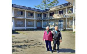 Two elderly people stand in the cement courtyard or front yard of a two-story housing complex.