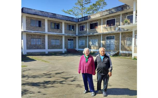 Two elderly people stand in the cement courtyard or front yard of a two-story housing complex.