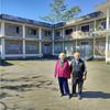 Two elderly people stand in the cement courtyard or front yard of a two-story housing complex.