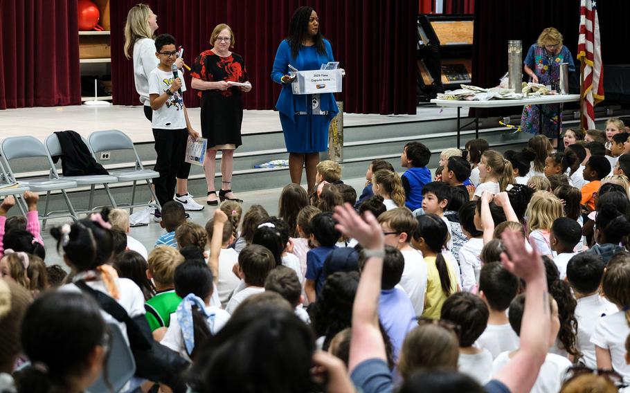 A crowd of seated elementary students watches as a student holding a microphone and adults present items.