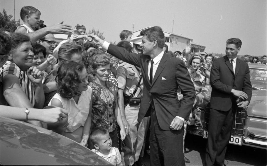 Fans circle RFK, who is extending his hand outward to shake a child’s hand.