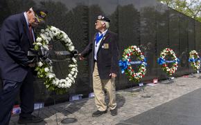 Two men in suits set up a wreath on a stand in front of a black memorial wall engraved with names as other wreaths line the wall and two other people approach carrying a wreath.