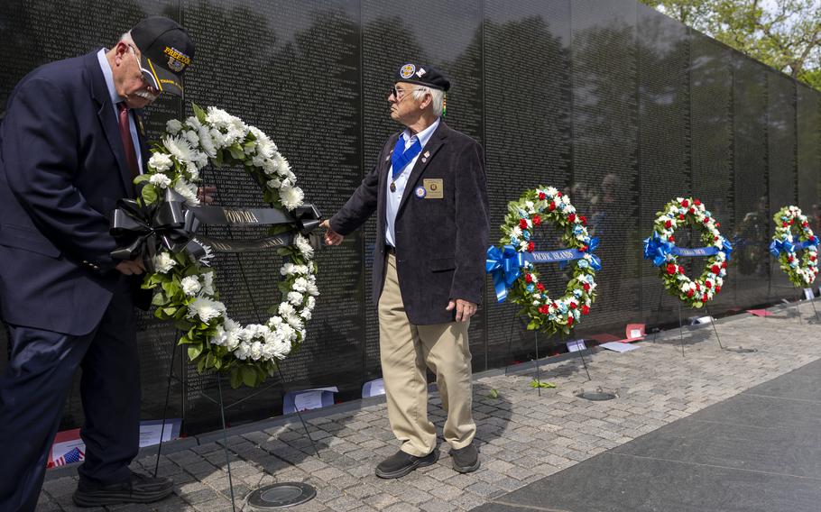 Two men in suits set up a wreath on a stand in front of a black memorial wall engraved with names as other wreaths line the wall and two other people approach carrying a wreath.
