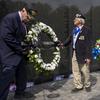 Two men in suits set up a wreath on a stand in front of a black memorial wall engraved with names as other wreaths line the wall and two other people approach carrying a wreath.