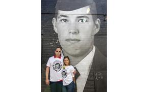 A man and a woman in white T-shirts stand with their arms around each other in front of a large portrait of an Air Force member in dress uniform.