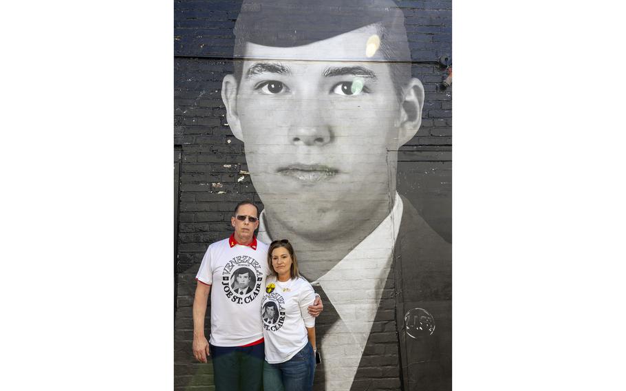 A man and a woman in white T-shirts stand with their arms around each other in front of a large portrait of an Air Force member in dress uniform.