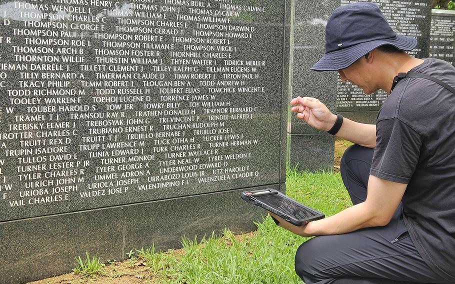 A man wearing gray shirt, pants and hat kneels on a patch of grass holding a tablet in front of a dark marble monument with lines of names engraved in white.