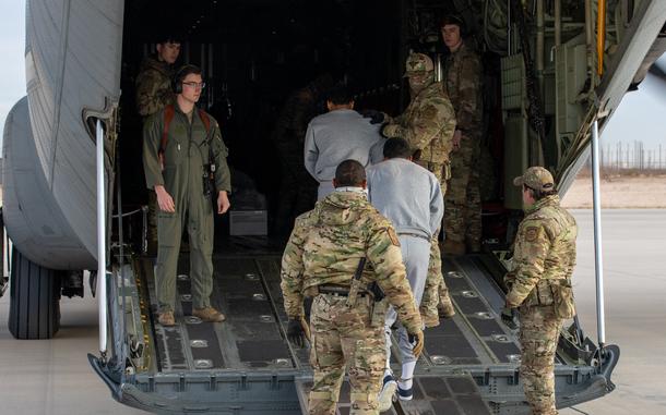 U.S. troops in camouflage uniforms guide detainees in gray sweatsuits up a ramp into a military aircraft on a runway.