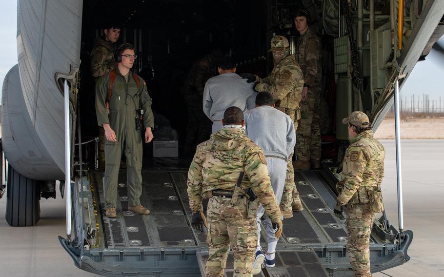 U.S. troops in camouflage uniforms guide detainees in gray sweatsuits up a ramp into a military aircraft on a runway.