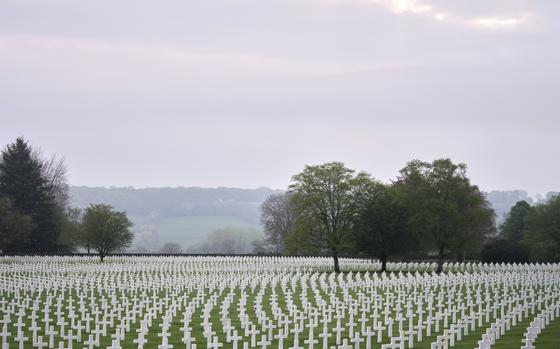 A vast military cemetery stretches across a green field, with hundreds of rows of white crosses marking graves. Trees with fresh spring leaves are scattered among the headstones, and a misty, overcast sky hangs overhead. Rolling hills are visible in the distance, adding to the solemn and peaceful atmosphere of the scene.