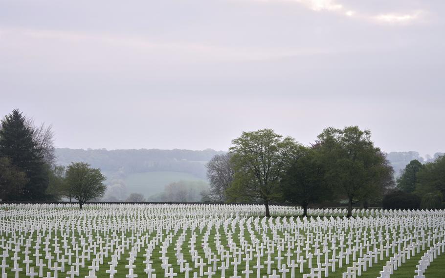 A vast military cemetery stretches across a green field, with hundreds of rows of white crosses marking graves. Trees with fresh spring leaves are scattered among the headstones, and a misty, overcast sky hangs overhead. Rolling hills are visible in the distance, adding to the solemn and peaceful atmosphere of the scene.