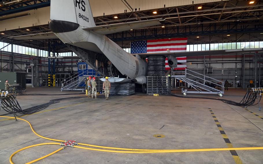 Rear view of a military transport aircraft with tail number “RS 84020” in a hangar. The aircraft’s cargo ramp is lowered and supported by numerous black inflatable air bags. Military personnel in camouflage uniforms stand near the aircraft. A large American flag hangs prominently in the background, and yellow testing cables snake across the hangar floor.