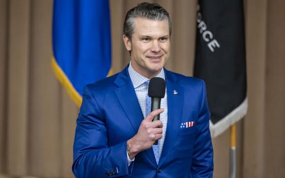 Pete Hegseth, wearing a blue suit, stands on a stage and holds a microphone.