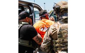A law enforcement agent in a black shirt and bulletproof vest, seen from behind, handcuffs a man in an orange shirt as a soldier in camouflage combat gear watches.