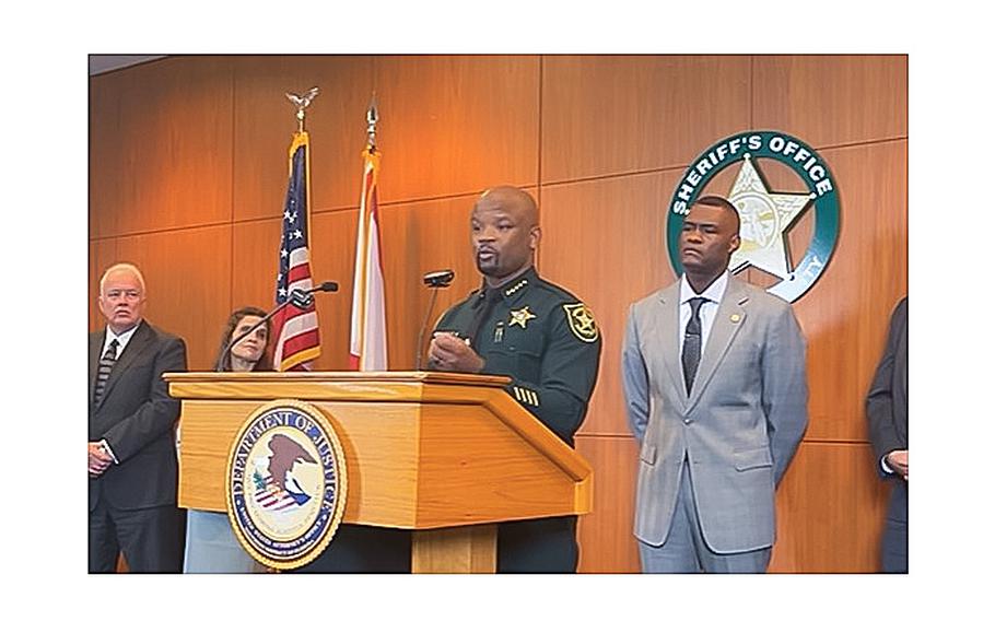 A video screen grab shows Broward County Sheriff Gregory Tony speaking at the podium as U.S. Attorney for the Southern District of Florida Markenzy Lapointe, back right, and other federal prosecutors announce on Thursday, Oct. 12, 2023, fraud charges against 17 officers related to allegations of stealing nearly $500K in covid-relief funds. 