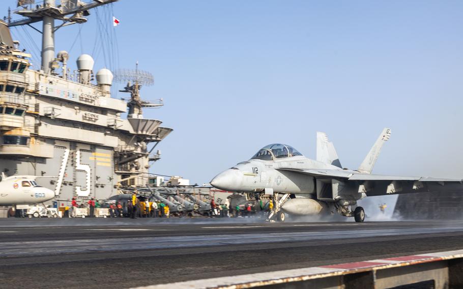 A military fighter jet speeds down the flight line as it takes off from an aircraft carrier at sea.