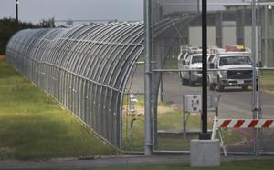 Two white trucks drive on a road behind the chain link fence of a government detention facility.